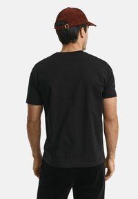 Man in een effen zwart T-shirt met korte mouwen en donkere broek, met zijn rug naar de camera gekeerd, en een bruine pet op zijn hoofd tegen een witte achtergrond.