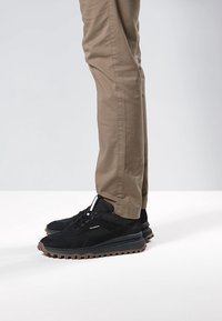 Schwarze Wildleder-Sneakers mit einer dicken Gummisohle und braunen Akzenten, kombiniert mit beigefarbenen Chinos, die ein lässiges Schuhdesign zeigen.