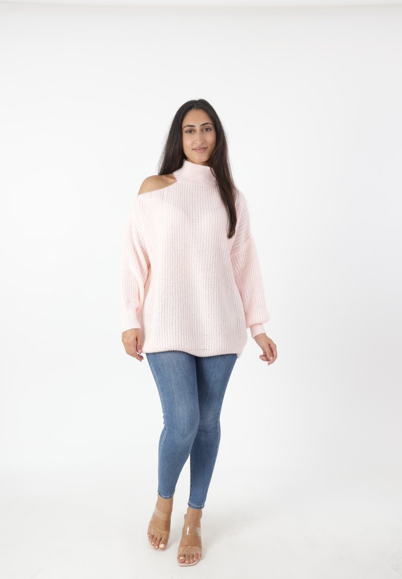 Maglione lavorato a maglia rosa chiaro con collo alto e un taglio sulla spalla, abbinato a jeans skinny blu e sandali con cinturini trasparenti.