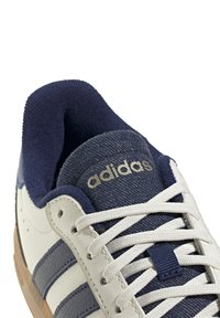 White sneaker with navy accents, featuring three stripes, textured fabric, and a rubber sole. "Adidas" logo embossed on the tongue.