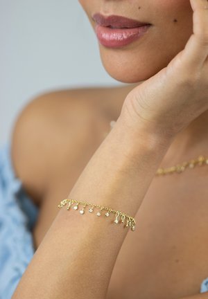 Woman's wrist with delicate gold bracelet featuring small dangling charms, close-up on skin and lips, light blue off-shoulder top visible.