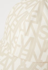 Beige sapka színazonos, ismétlődő szövegmintával. Simaságával és strukturált paneleivel, valamint ívelt karimájával tűnik ki. Öltözködési részletekkel van kiemelve.