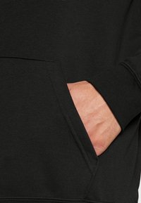 Primo piano di un tessuto di felpa nera che mostra una mano nella tasca frontale, caratterizzato da una texture liscia e polsini elasticizzati.