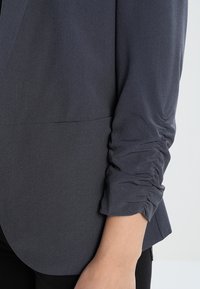 Mörkgrå tygblazer med en rynkad ärmdesign och en slät yta. Skräddarsydd passform med rundad kant vid fållen.