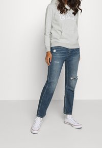 Sudadera gris con el logo "GAP", jeans azules desgastados con rotos y zapatillas altas blancas. Conjunto casual presentado por un modelo.