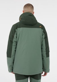 Grüne isolierte Jacke mit hohem Kragen, kontrastierenden dunkeleren Ärmeln und Logodetail. Glatte Textur mit Kapuze und verstellbaren Bündchen.