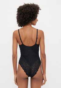 Fekete csipkebody vékony, állítható pántokkal, virágmintával, fodros szélekkel és testhezálló fazonnal, amely a sima sziluettet hangsúlyozza.