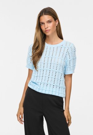 Jeune femme aux longs cheveux bruns portant un haut en maille bleu clair à manches courtes et un pantalon noir, debout devant un fond uni.