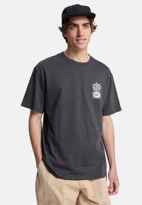 Γκρι βαμβακερό μπλουζάκι με κοντά μανίκια, με λευκό γραφικό σχέδιο στο αριστερό μέρος του στήθους. Συνδυασμένο με μπεζ σορτς.