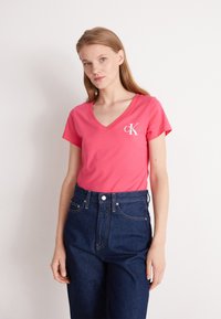 Camiseta de algodón rosa con escote en V y un logo blanco en el pecho izquierdo, combinada con jeans oscuros de tiro alto.