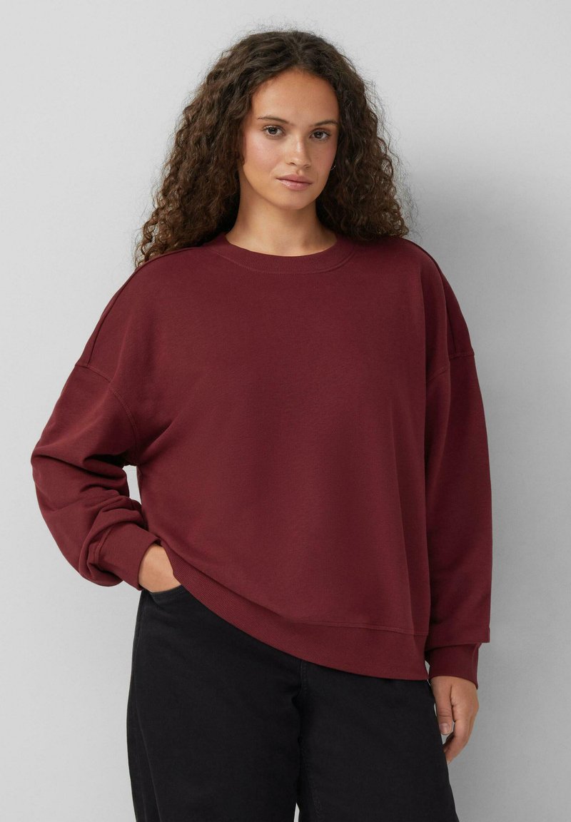 Jonge vrouw met krullend bruin haar die een losse bordeauxrode sweater en zwarte broek draagt, staand met één hand in de zak tegen een grijze muur.