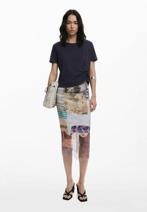 Dunkelblaues T-Shirt mit geknoteter Vorderseite, kombiniert mit einem mehrfarbigen, transparenten Rock mit verschiedenen Texturen und Farben. Schwarze, riemchen Sandalen.