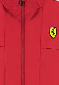 Czerwona kurtka z suwakiem z przodu, z czarno-żółtym logo Ferrari. Gładka tekstura, standardowy kołnierz i dwie boczne kieszenie.