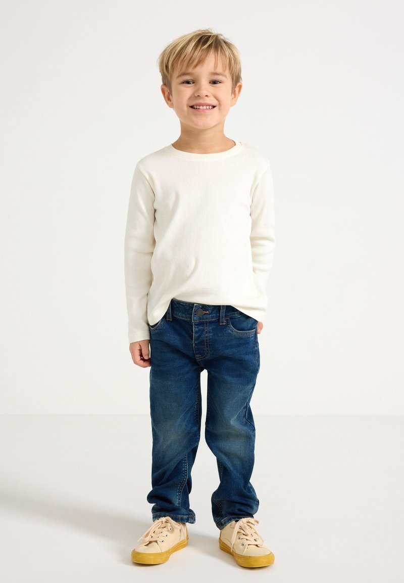 Maglietta bianca a maniche lunghe a coste, jeans in denim blu e sneakers beige. La maglietta ha un collo rotondo, i jeans presentano tasche standard e passanti per cintura.