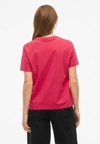 Koszula z krótkim rękawem z żywej różowej bawełny, z okrągłym dekoltem i luźnym krojem, w zestawieniu z czarnymi spodniami. Widok od tyłu.