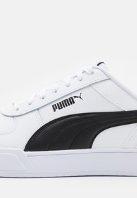 Sneaker bianco con una striscia laterale in pelle nera, lacci bianchi e logo Puma nero. Texture liscia con design low-top.