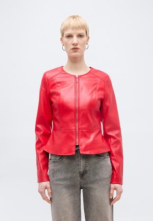 Vrouw met kort blond haar draagt een rode leren jas met rits en grijze jeans, staand tegen een effen lichte achtergrond.