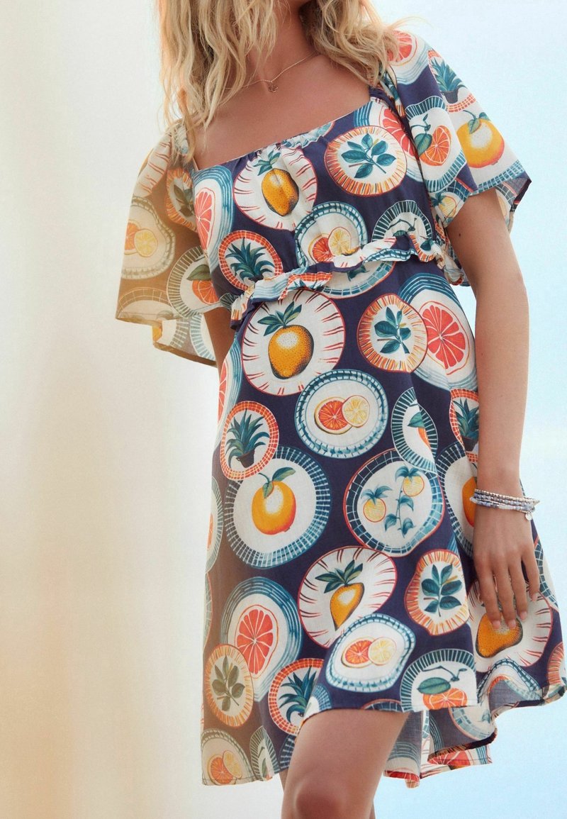 Vestido de manga corta con un vibrante estampado de frutas en patrones circulares, con fondo azul marino, cintura fruncida y tejido ligero.