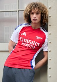 Tricou sportiv roșu și alb cu accente albastre, având imprimat "Emirates Fly Better" și un design cu trei dungi pe mâneci, asortat cu pantaloni gri.
