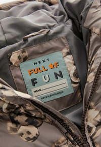 L'etichetta all'interno di una giacca grigia e con motivo riporta "NEXT FULL OF FUN" con una linea per il nome vuota e "Fatto in Bangladesh."