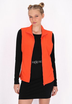 Orange fleece vest with a high collar and front zip. Worn over a long-sleeve black dress, accentuated by a black belt.