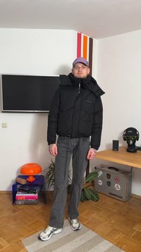 Svart dunjakke med glidelås og hette, brukt over gråvidde jeans med sleng. Bakgrunnen har en TV og dekorative gjenstander.