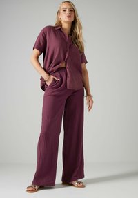 Completo a due pezzi in tessuto testurizzato di un viola scuro. Camicia a maniche corte con bottoni abbinata a pantaloni larghi con vita elastica. Sandali piatti.