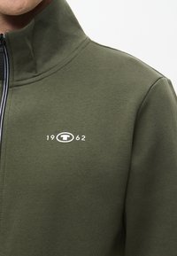 Sötétzöld, cipzáros dzseki puha anyagból, magas nyakkal és a mellkasán elhelyezett, fehér "1962" logóval.