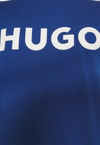 Tissu bleu avec de grandes lettres blanches en gras formant "HUGO" sur toute la surface.