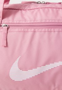 Rózsaszín anyagú hátizsák sima felülettel, fehér Nike swoosh logóval és cipzáras zsebekkel.