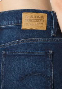 Džínové kalhoty s tmavě modrým odstínem, hnědou koženou nášivkou s embosovaným textem a jemnými švy na zadní kapse.