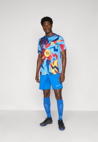 Rövid ujjas kék sportpóló színes absztrakt mintákkal, kék rövidnadrággal és fekete focicipőkkel párosítva. Logó részletekkel rendelkezik.