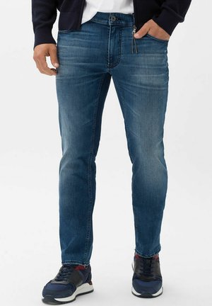 Dunkelblaue Jeans im Slim-Fit-Stil, mit subtilen Verwaschungen und einem klassischen Fünf-Taschen-Design. Kombiniert mit dunkelblauen Sportschuhen.