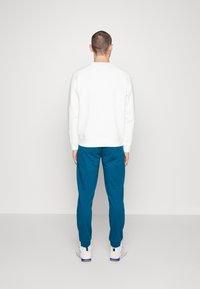 Man som står vänd bort med en vit sweatshirt, turkosa joggarbyxor och vita sneakers mot en enkel vit bakgrund.