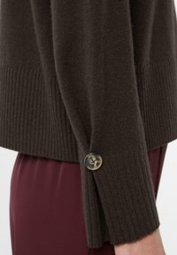 Dunkelbrauner Strickpullover mit gerippter Textur und einem einzelnen Knopf-Detail am Bündchen. Verfügt über eine lockere Passform und weiches Material.