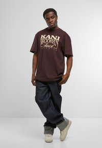 Dunkelbraunes, übergroßes T-Shirt mit cremefarbenem Grafiktext. Kombiniert mit weit geschnittenen, dunkelblauen Jeans und hellen Sneakers mit umgeschlagenem Saum.