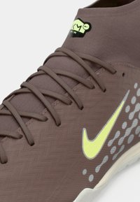 Brūnas sporta kurpes ar teksturētu materiālu, ar dzeltenu Nike swoosh, pelēkiem akcenta rakstiem un brūnām šņorēm.