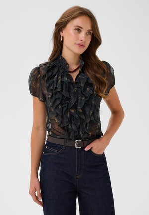 Kvinde med langt brunt hår iført en gennemsigtig sort bluse med flæser, mørke højtaljede jeans, et bælte og en bordeauxrød perlekæde.