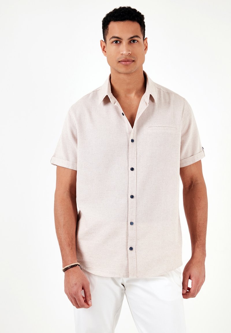 Camisa de manga corta de color beige claro con tejido texturizado, botones azules y un bolsillo en el pecho. Presenta mangas remangadas y un dobladillo curvado.