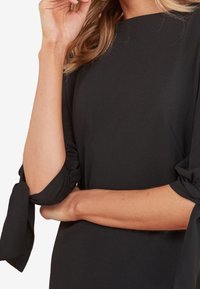 Femme portant une robe noire à manches longues avec des détails en nœud noués sur les manches, tenant un bras sur sa taille et touchant son menton.