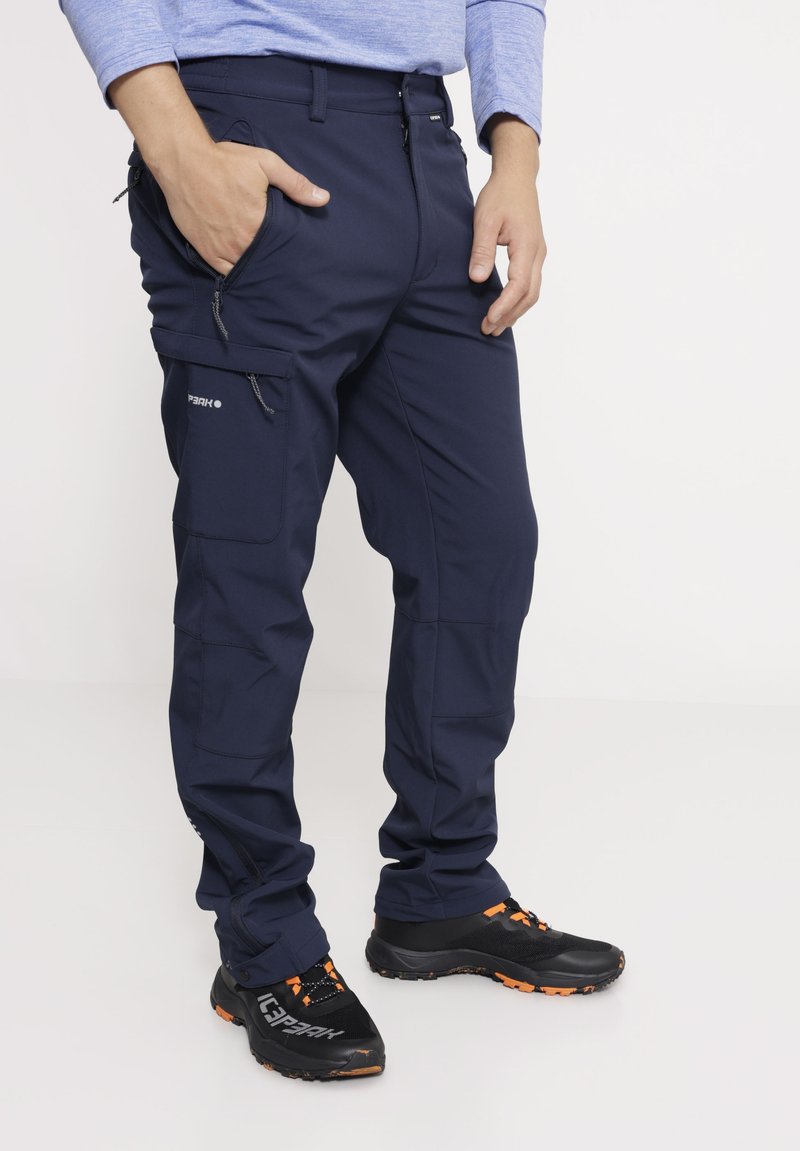 Dunkelblaue Outdoor-Hosen aus strapazierfähigem Material, mit Reißverschlusstaschen und einem lässigen Schnitt. Kombiniert mit schwarzen Schuhen mit orangefarbenen Akzenten.