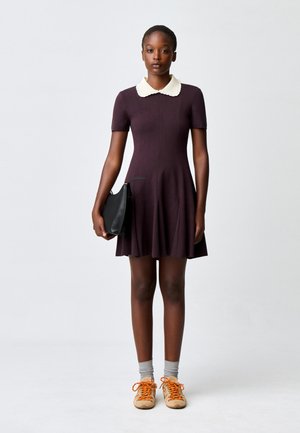 Giovane donna che indossa un vestito scuro a maniche corte con colletto bianco, tiene una borsa nera, calzini grigi e sneakers beige con lacci arancioni.