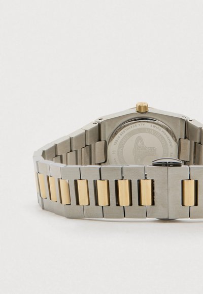 Bracelete de relógio em metal prateado e dourado com fecho, mostrando o verso da caixa do relógio com o nome da marca gravado e detalhes do design.