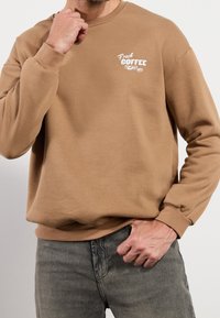 Sudadera de algodón marrón con cuello redondo y puños acanalados. Presenta texto blanco "Fresh Coffee" con un gráfico de granos de café en el pecho.