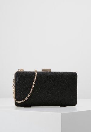 Pochette rectangulaire noire à texture pailletée avec une bandoulière chaîne dorée, posée sur une surface blanche devant un fond gris clair.