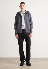 Sudadera gris con cremallera y capucha, mangas largas y bolsillos frontales, llevada sobre una polo gris claro con detalles negros y pantalones negros.