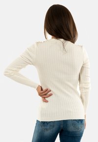 Maglione bianco lavorato a maglia con un design testurizzato e a coste; presenta dettagli sulle spalle e maniche aderenti. Indossato con jeans blu.