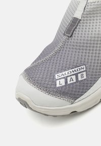 Szürke hálós sportcipő texturált felső résszel, fehér részletekkel és "SALOMON LAB" márkával az oldalon. Lekerekített orrforma.