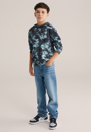 Gezicht op een hoodie met een zwart en teal tie-dye patroon, gecombineerd met lichtblauwe spijkerbroek en zwarte hoge sneakers met witte accenten.