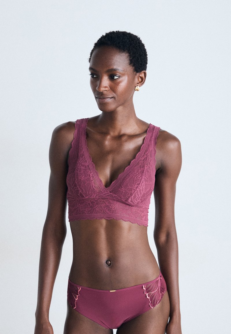 Kvinne i matchende mørk rosa blonde bralette og broderte truser, som står mot en ensfarget lys bakgrunn og ser til venstre.